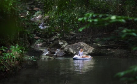 Mattress upon a flowing stream in lush green rainforest with woman meditating on float mattress, peaceful nature scene, outdoor relaxation, natural wellness, tranquil water body, eco-friendly lifestyle, mind-body harmony.