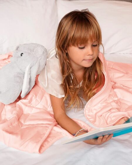 Soft pink comforter on a bed with a plush bunny toy, young girl reading a book.